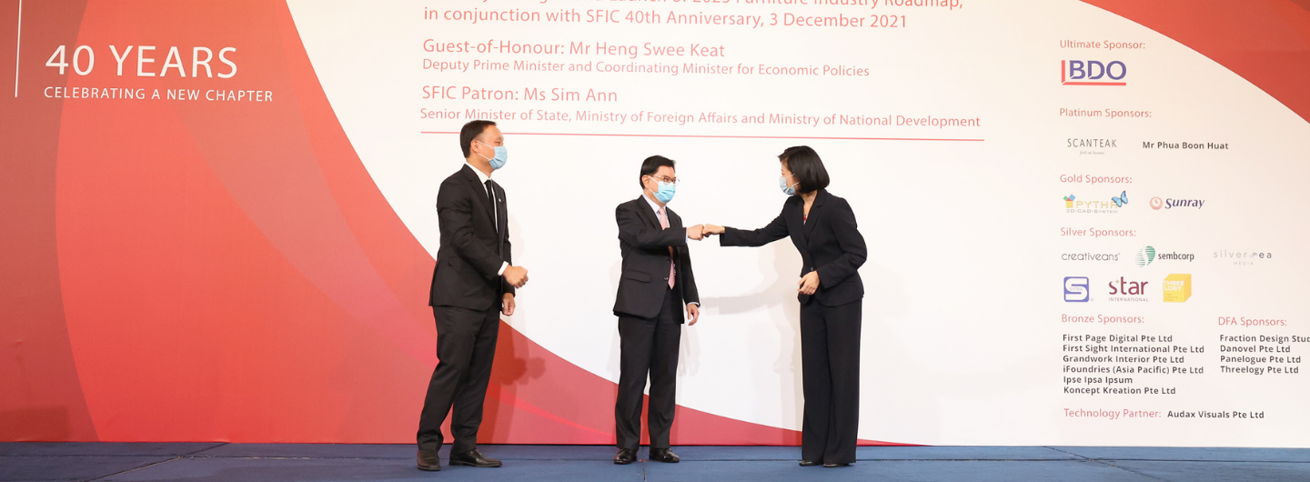 Three people in suits and face masks touch fists in front of event signage listing sponsors like BDO.